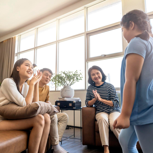 Family sitting together in a living room, smiling and clapping as a young girl stands in front of them, suggesting support, encouragement, and positive family connection.
