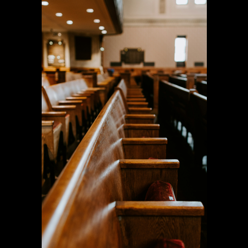 Rows of wooden benches inside an empty courtroom, viewed from the side with soft lighting and a blurred judge’s bench in the background.